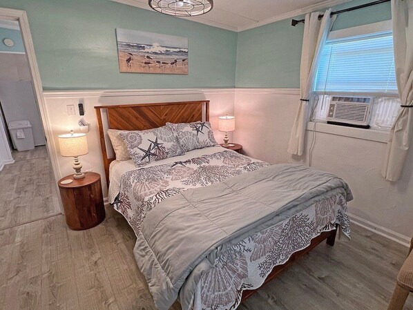 A bedroom with a bed, two bedside tables, a wall-mounted heater, and a window with blinds.