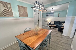 A dining area with a wooden table and chairs, a hanging light fixture, and a view into the living room with a sofa and a lamp.