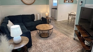 A living room with a dark blue sofa, a round wooden coffee table, and a patterned rug. There is a TV stand with a flat-screen TV and a lamp on a side table. - Auralee Unit 1 - Beach Access - Island Realty (Grand Isle)