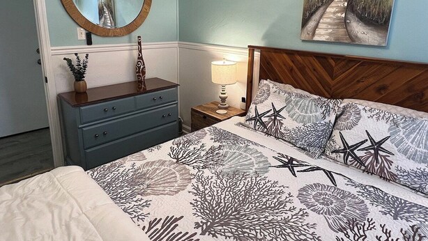 A bedroom with a bed, a dresser, a nightstand, a lamp, a mirror, and a painting on the wall. - Auralee Unit 2 - Beach Access - Island Realty (Grand Isle)