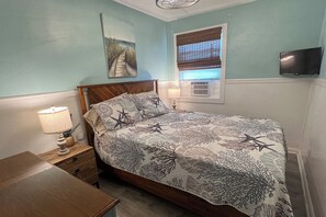 A bedroom with a bed, a nightstand, a lamp, a painting on the wall, a window with blinds, and an air conditioning unit. - Auralee Unit 2 - Beach Access - Island Realty (Grand Isle)