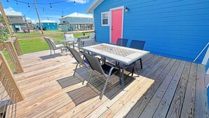 En træterrasse med udendørs møbler, en blå bygning med en lyserød dør og en klar himmel.