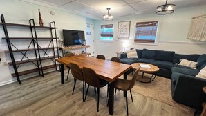A modern living room with a wooden dining table, a sofa, and a television. - Auralee Unit 2 - Beach Access - Island Realty (Grand Isle)