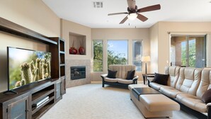Un salon moderne doté d’un téléviseur à écran plat, d’un ventilateur de plafond, d’une cheminée, d’un canapé et d’une grande fenêtre offrant une vue sur les arbres.