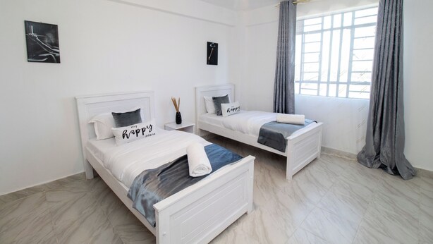 Een moderne slaapkamer met twee eenpersoonsbedden, witte hoofdborden en een groot raam met grijze gordijnen.