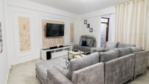 Een woonkamer met een grijze bank, een flatscreen televisie aan de muur, een witte dressoarkast en een grijze accent fauteuil.