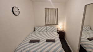 Una camera piccola con un letto singolo, una coperta a righe, un comodino con una lampada, una sveglia a parete e una finestra con tende.