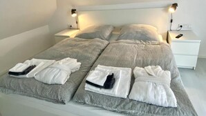 A neatly made bed with grey bedding and white towels, two bedside tables with lamps, and a white nightstand with a phone on it. - Spacious modern DG apartment Schloofschdubb near Kassel 70 sqm (Fuldatal)