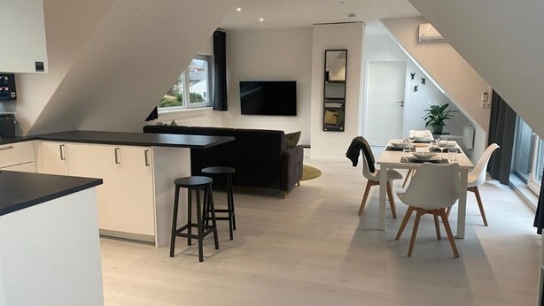 A modern kitchen with a central island, bar stools, a dining area with white chairs, and a flat-screen TV mounted on the wall. - Spacious modern DG apartment Schloofschdubb near Kassel 70 sqm (Fuldatal)