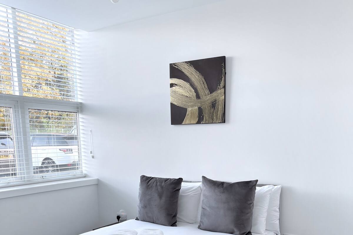 Un letto rifatto con lenzuola bianche e cuscini grigi, un comodino nero con una lampada e un quadro appeso al muro.