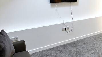 Una sala de estar moderna con una televisión de pantalla plana montada en la pared, una alfombra gris y un sofá gris con un cojín oscuro.