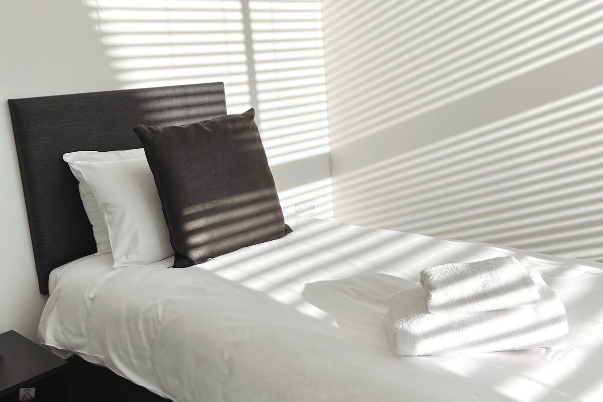Un letto rifatto con lenzuola bianche e due cuscini, una testiera scura e un comodino con una lampada.
