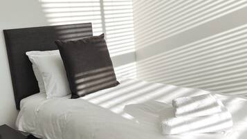Una cama bien tendida con sábanas blancas y dos almohadas, un cabecero oscuro y una mesita de noche con una lámpara.