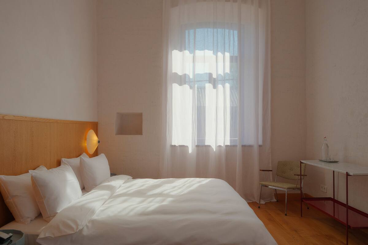 A hotel room with a bed, a chair, a small table, and a window with sheer curtains.