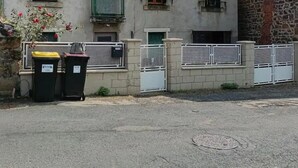 Un bâtiment à deux étages, doté d’une façade en pierre et de nombreuses fenêtres avec des volets. On y trouve deux poubelles et une entrée fermée par un portail.