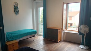 Une pièce comprenant un lit, une commode, une fenêtre avec des rideaux et un ventilateur sur pied.