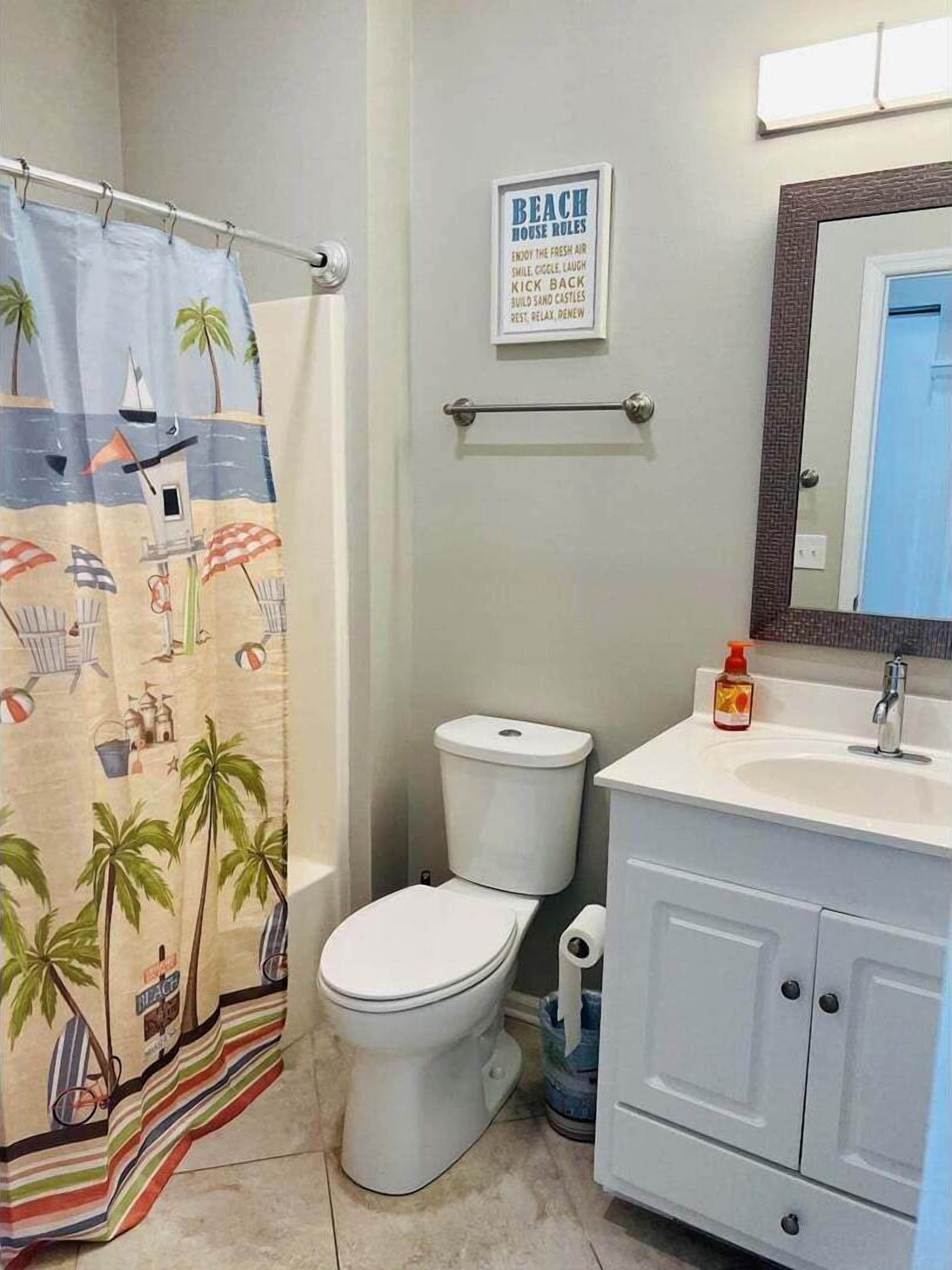 A bathroom with a toilet, a sink, a shower curtain with a beach theme, and a framed sign with beach house rules.