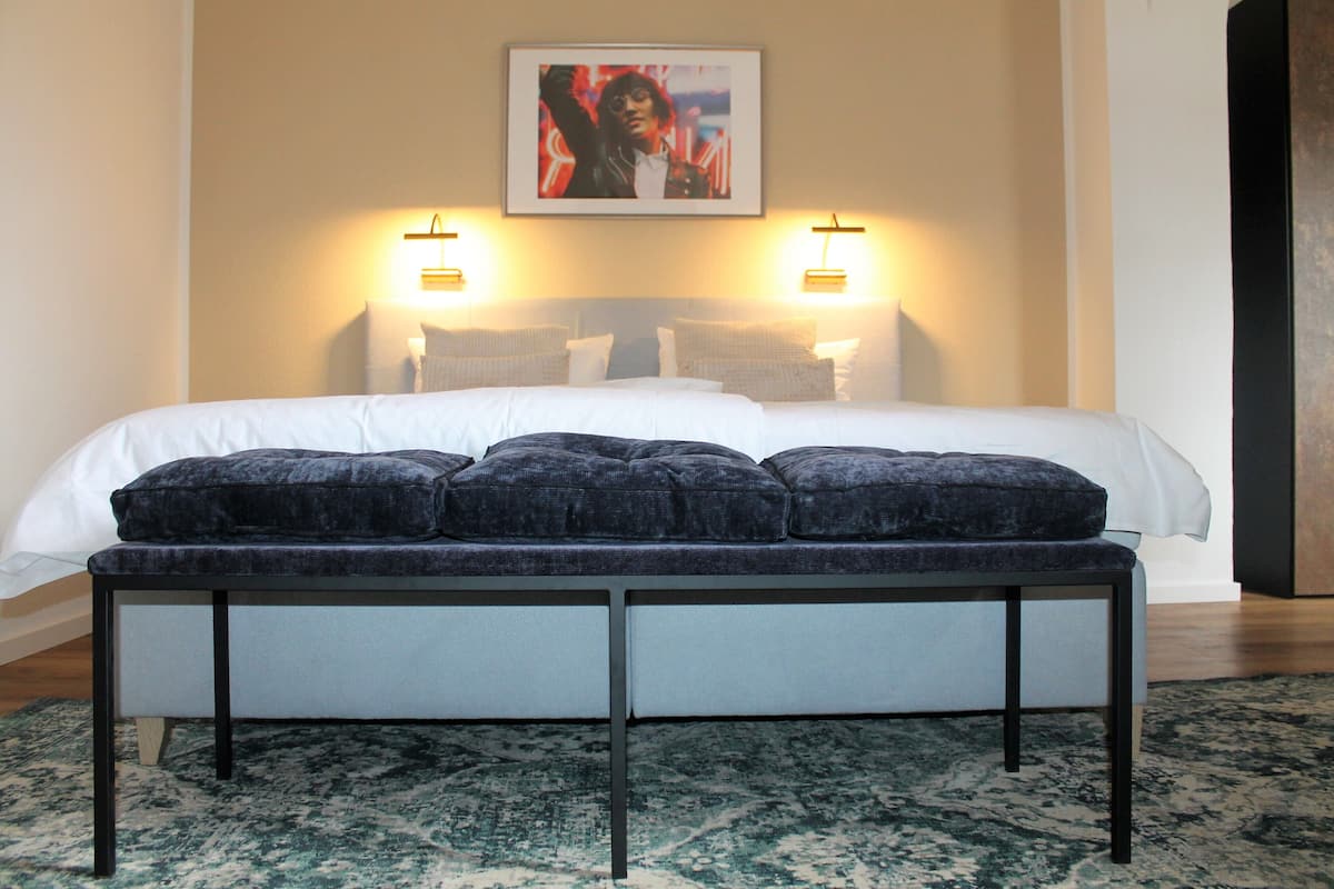 A neatly made bed with a white duvet and dark blue pillows, a bench with a cushion, and two wall-mounted lamps.