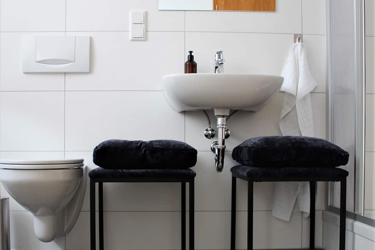 A modern bathroom with a white sink, two black stools, a toilet, and a mirror above the sink.