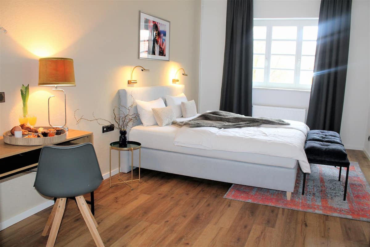 A modern bedroom with a white bed, a desk with a lamp, a chair, and a window with curtains.