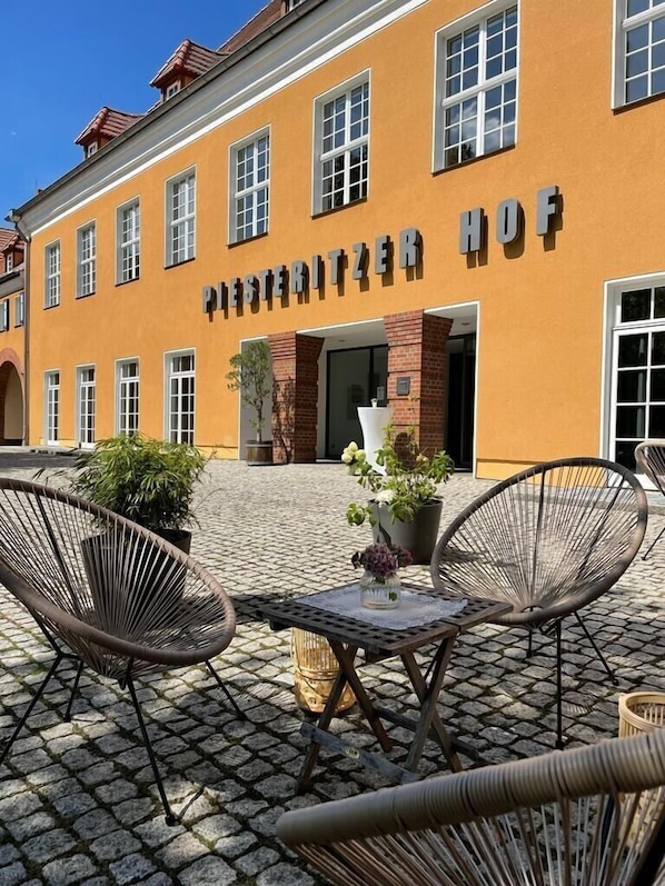Exterior de hotel con fachada amarilla, múltiples ventanas y zona de estar al aire libre con sillas de mimbre y una mesa.
