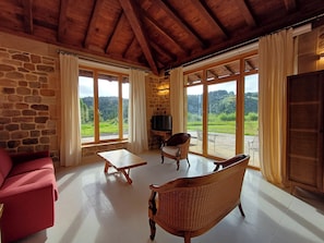 A room with stone walls, wooden ceiling beams, a red sofa, a wicker chair, a small table, and a TV. - New apartment with garden in a rural house with sea views (Vizcaya)
