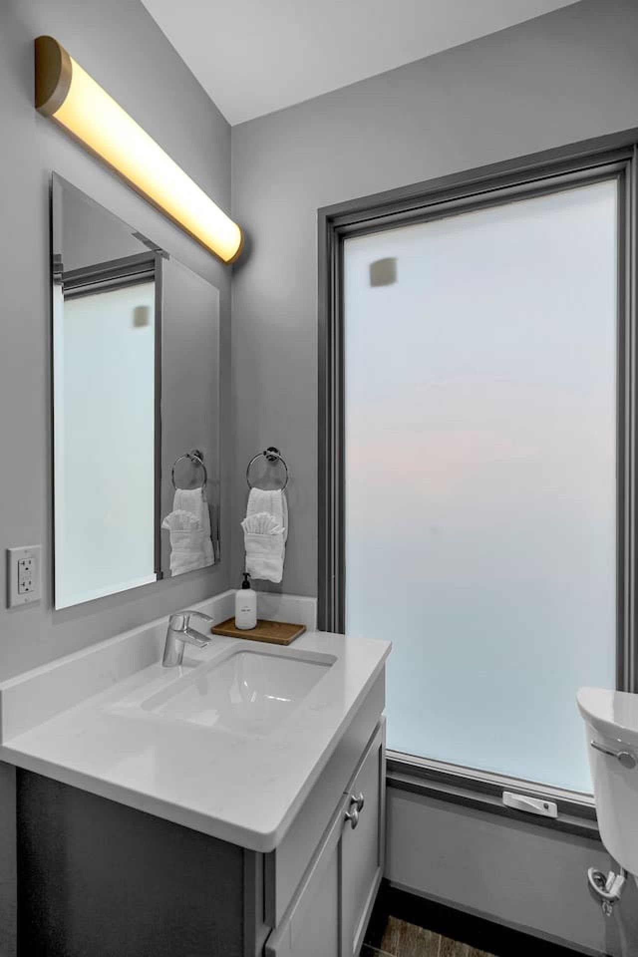 Une salle de bain moderne avec un grand miroir, un lavabo blanc et des toilettes. Il y a une fenêtre avec du verre dépoli et un porte-serviettes avec deux serviettes blanches.