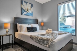Une chambre avec un grand lit, deux lampes de chevet, une fenêtre avec des stores et une œuvre d’art florale accrochée au mur.