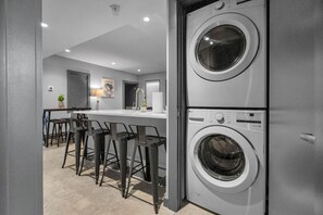 Un lavadero moderno con dos lavadoras apiladas, una zona de bar con taburetes y una puerta que da a otra habitación.