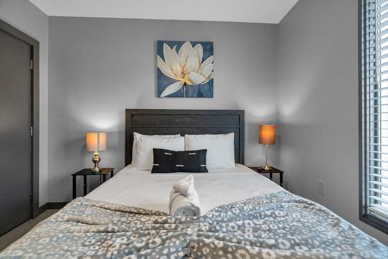 A hotel room with a large bed, two bedside lamps, a wall-mounted artwork of a flower, and a window with blinds.