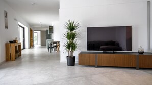 A modern living room with a large flat-screen TV, a wooden console table, a potted plant, and a dining area in the background.