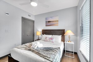 Une chambre d’hôtel bien rangée, avec un lit, deux lampes de chevet, un tableau et une fenêtre avec des stores.