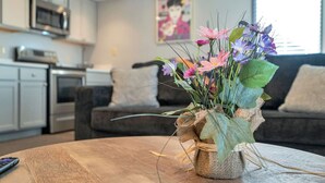 Un salon avec une table basse en bois, un vase de fleurs, un canapé et une cuisine en arrière-plan.
