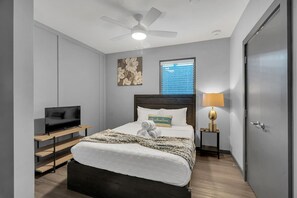 Een slaapkamer met een bed, een televisie, een plafondventilator, een raam met zonwering en een deur.