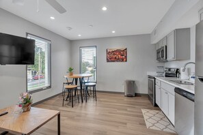 Een moderne keuken met witte kastjes, een magnetron en een vaatwasser. Er is een eethoek met een tafel en stoelen, en een televisie aan de muur bevestigd.