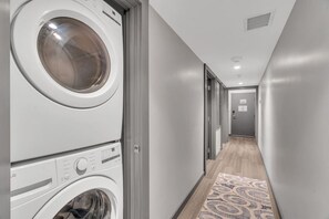 Une buanderie avec des lave-linge et des sèche-linge superposés, un tapis à motifs et un couloir menant à une porte.
