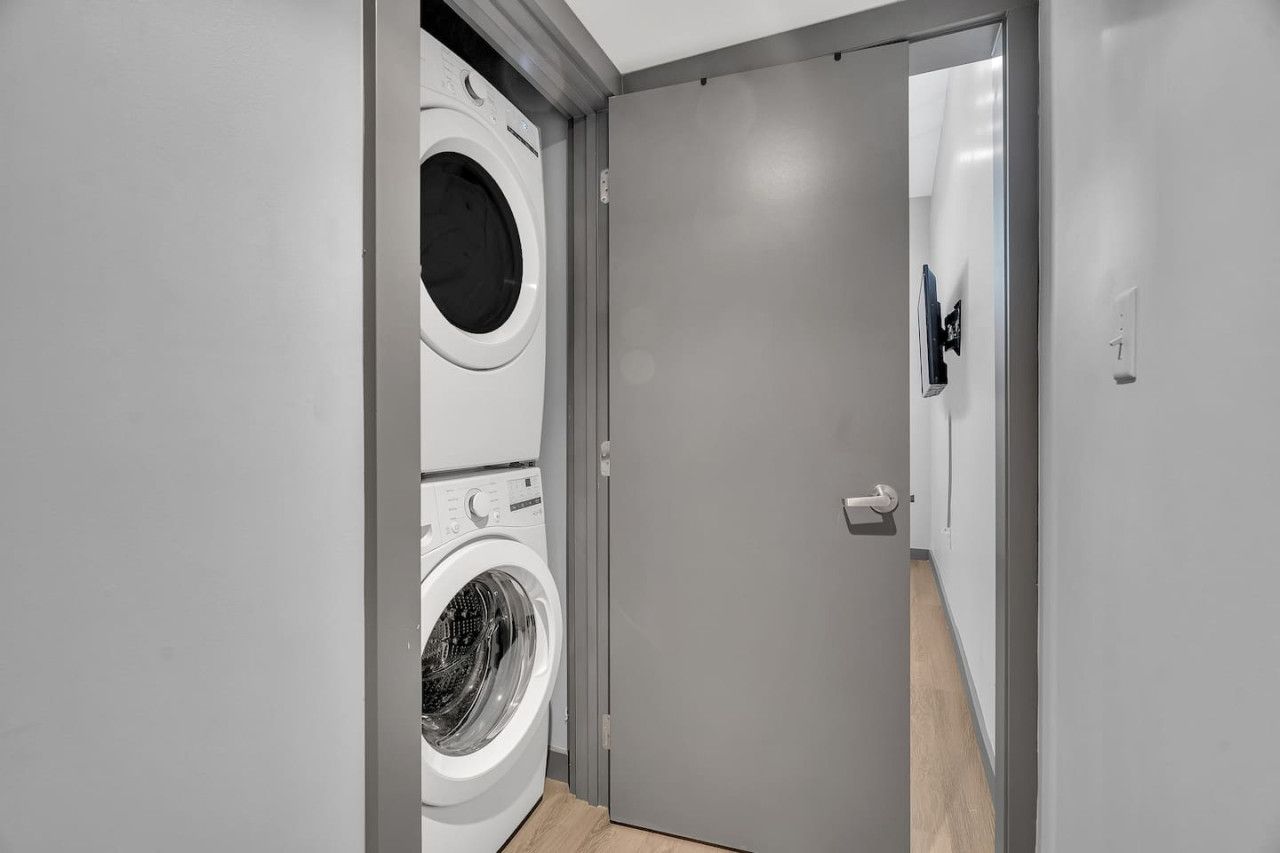 A modern laundry room with a front-loading washer and dryer, a grey door, and a visible light switch.