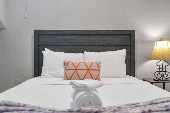 Un lit bien fait, avec des draps blancs, un coussin décoratif orange et une serviette blanche roulée posée sur le lit.
