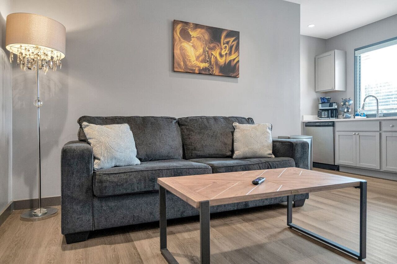 A modern living room with a grey sofa, a wooden coffee table, a floor lamp, and a kitchen area with cabinets and a window.