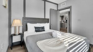 A modern bedroom with a bed, a nightstand, a lamp, and a view into the kitchen. - Cozy New Build Near FQ/Frenchman+Parking (New Orleans)