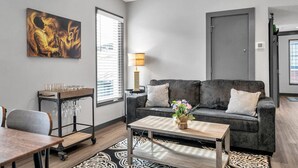A modern living room with a grey sofa, a coffee table, a dining table, and a wall-mounted artwork. - Cozy New Build Near FQ/Frenchman+Parking (New Orleans)