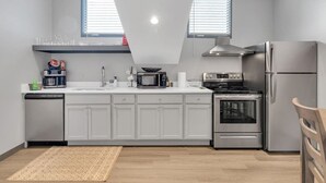 A modern kitchen with white cabinets, a stainless steel refrigerator, a microwave, a stove, and a dishwasher. There are two windows with blinds, a wooden chair, and a patterned rug on the floor. - Bywater Haven: Near FQ&Frenchman + Secure Parking (New Orleans)
