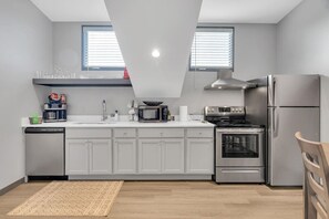 Une cuisine moderne avec des armoires blanches, un réfrigérateur en acier inoxydable, un four à micro-ondes, une cuisinière et un lave-vaisselle. Il y a deux fenêtres équipées de stores, une chaise en bois et un tapis à motifs sur le sol.