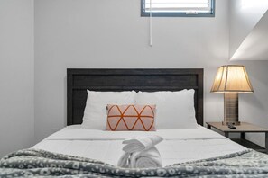 Un lit bien fait, agrémenté d’un coussin à motifs, d’une lampe de chevet et d’une fenêtre située au-dessus du lit.