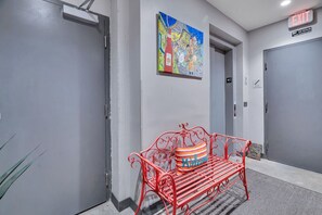 Un pasillo con puertas grises, un mural colorido y un banco rojo con un cojín que dice 'Welcome'.