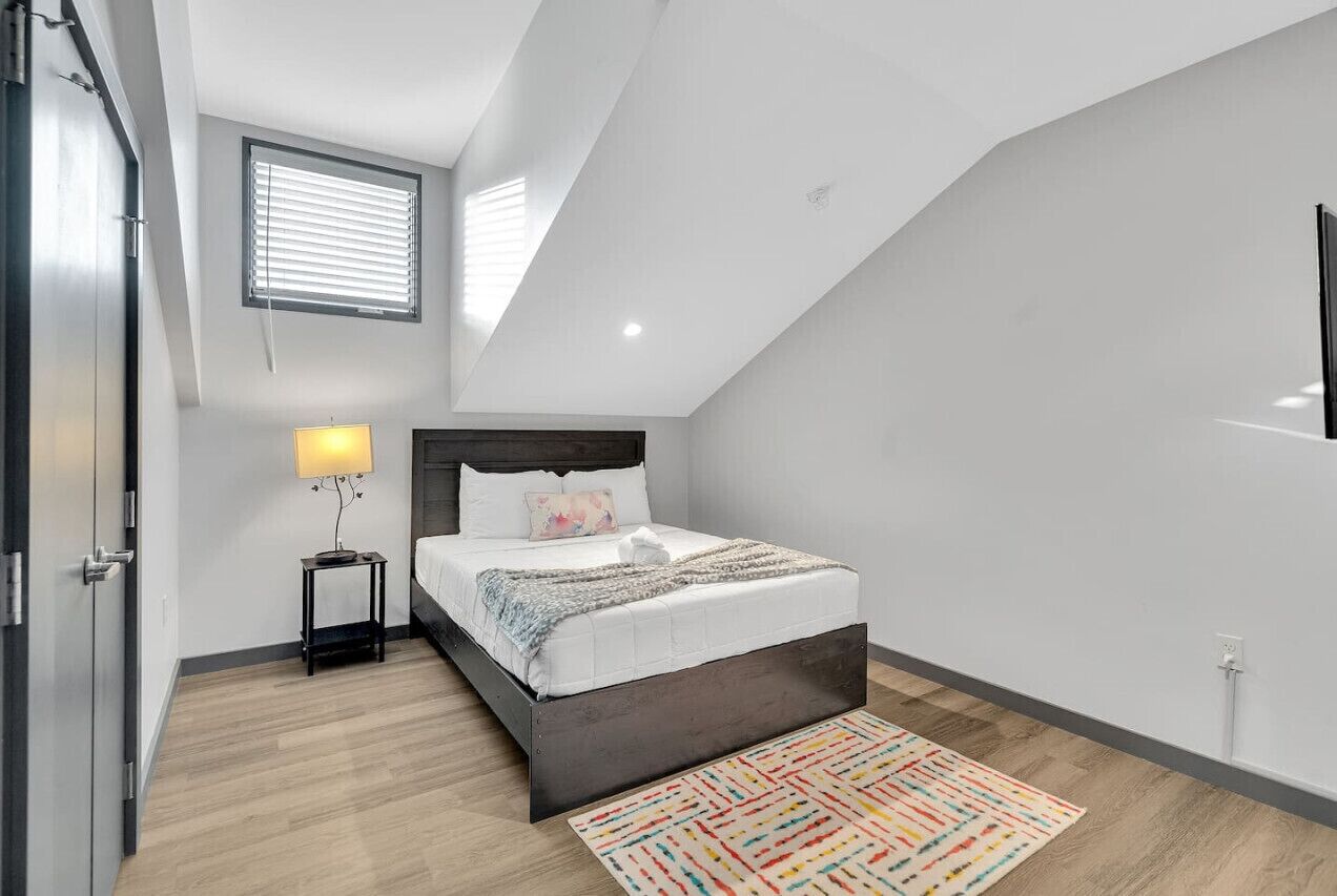Un dormitorio moderno con techo inclinado, una cama con ropa blanca, una mesita de noche con lámpara y una alfombra colorida.