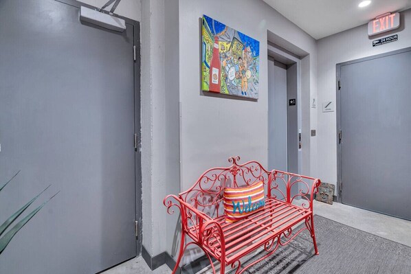 Un couloir moderne avec des portes grises, un banc rouge agrémenté d’un coussin sur lequel est écrit « Bienvenue », et une fresque colorée sur le mur.