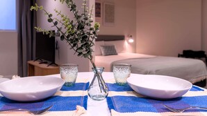 A hotel room with a dining table set for two, two bowls, two glasses, and a vase with a plant.
