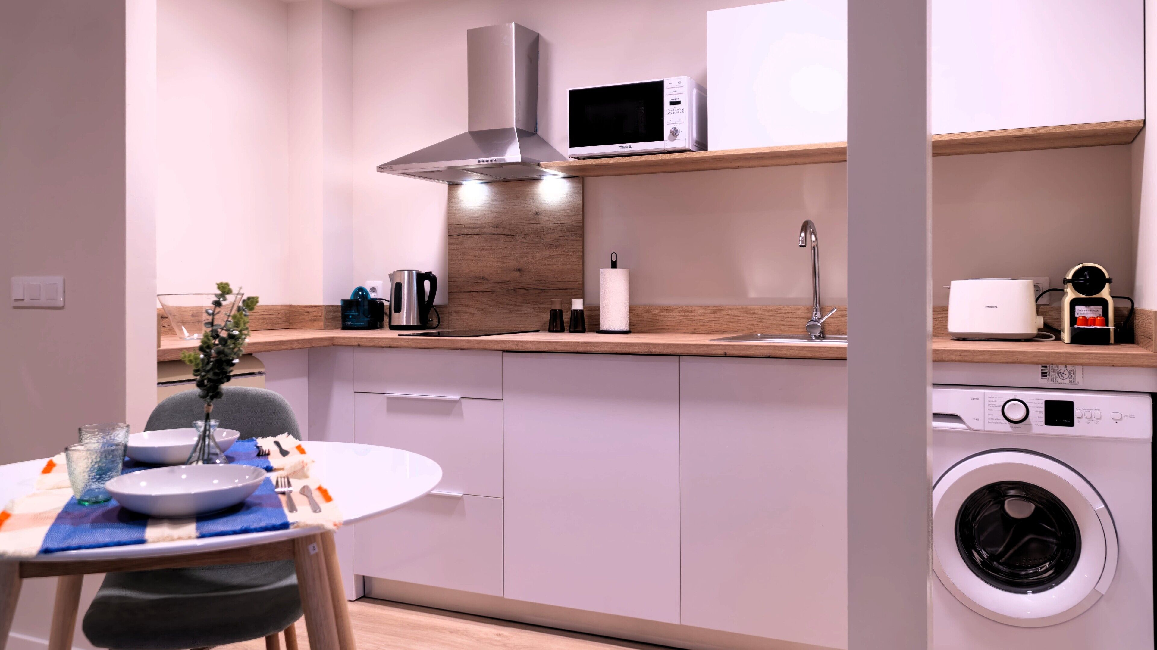 A compact kitchen with a microwave, kettle, and toaster on the countertop, a sink, and a washing machine.