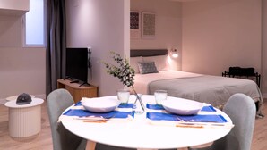 A modern hotel room with a neatly made bed, a dining table set for two, a television, and a small side table with a remote control.